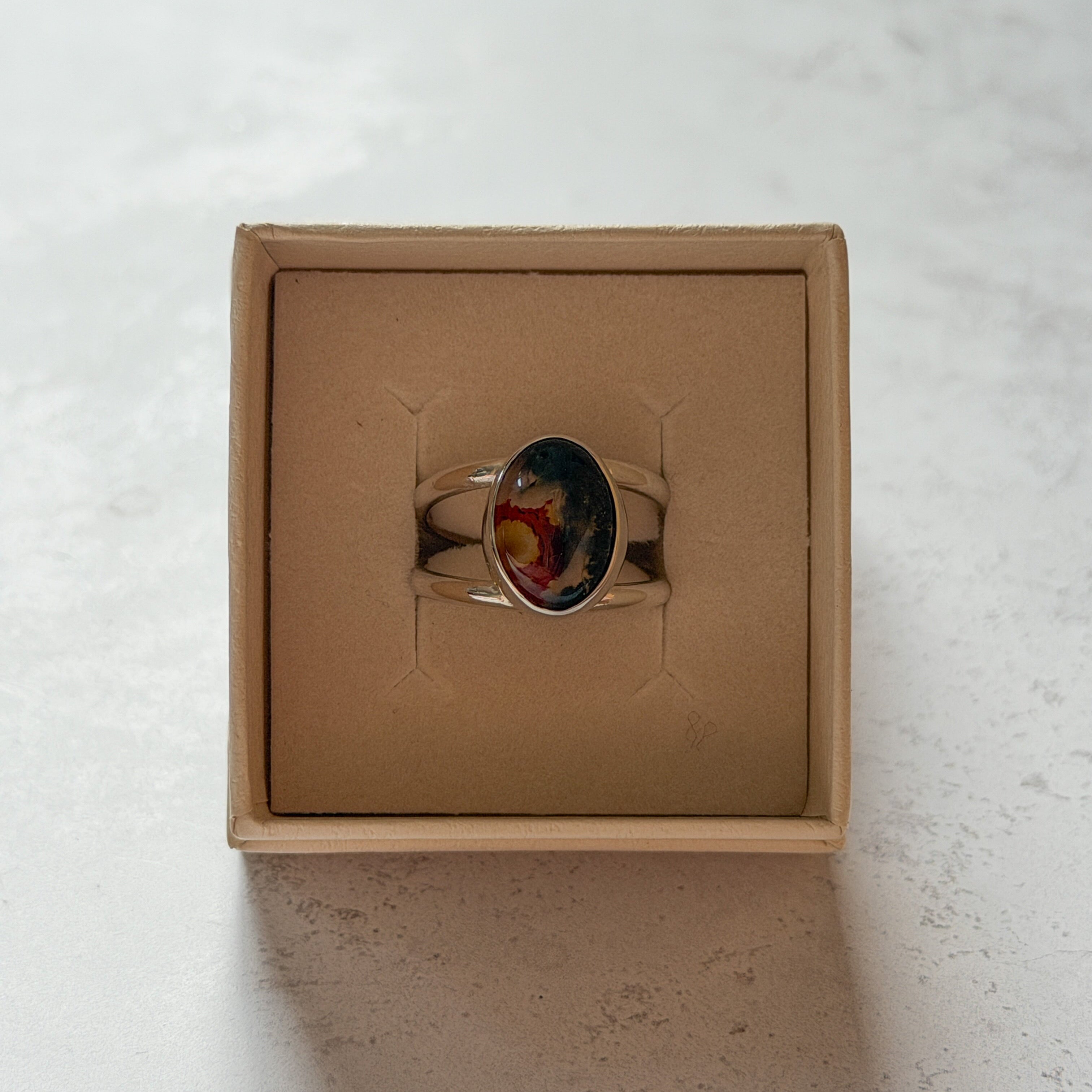 Handmade sterling silver double band ring featuring an oval moss agate and carnelian gemstone with a vivid natural red inclusion. One-of-a-kind artisan statement ring by Journey Through Jewellery, displayed in a cream jewellery box with a polished finish.