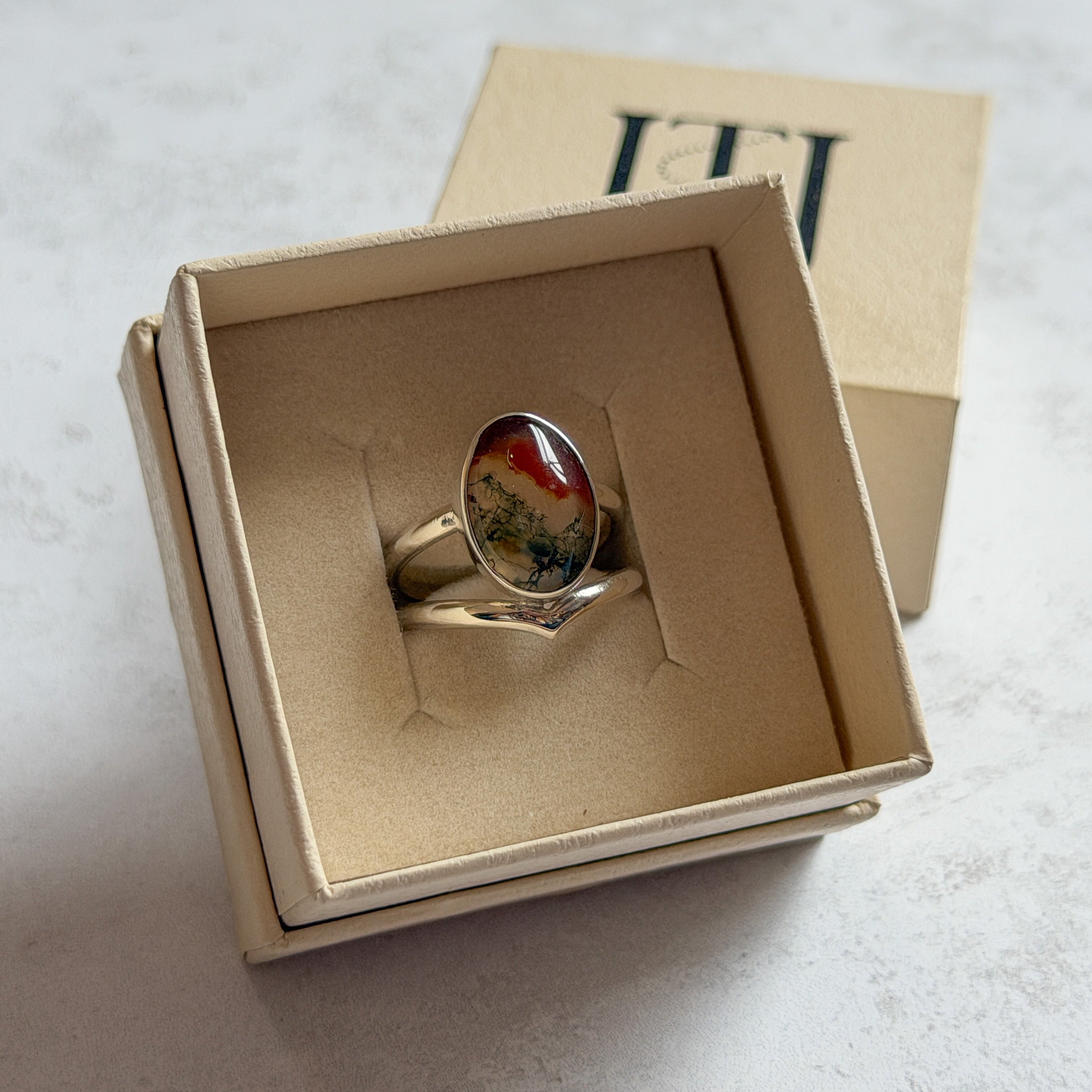 Pair of handcrafted sterling silver stud earrings with oval moss agate and carnelian gemstones, displayed in a cream Journey Through Jewellery gift box. Each one-of-a-kind gemstone features unique amber, green and black landscape-like inclusions, finished with mirror-polished bezels and handcrafted silver grain details.