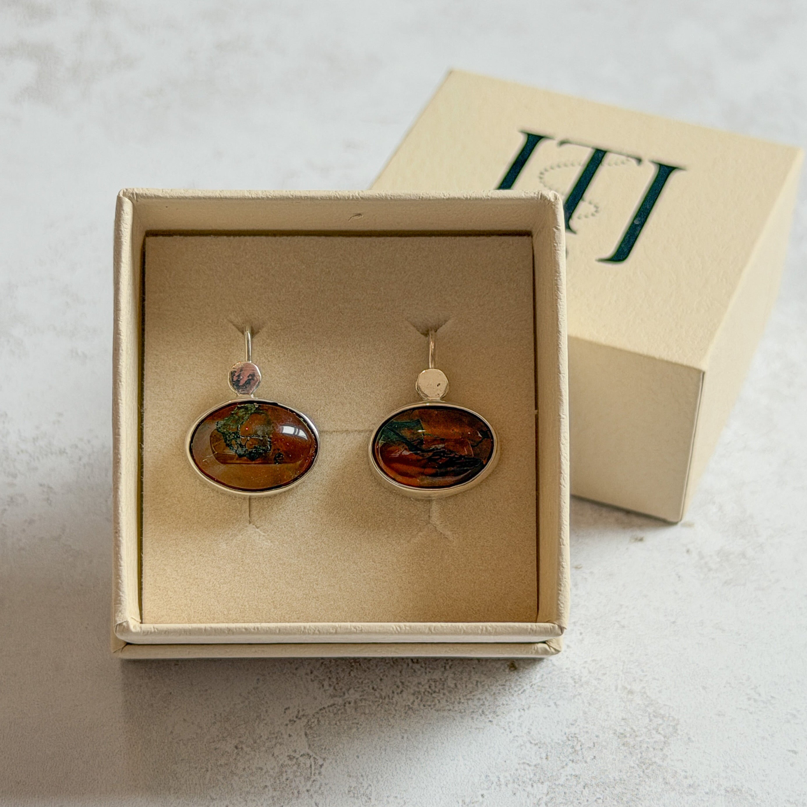 Pair of handcrafted sterling silver stud earrings with oval moss agate and carnelian gemstones, displayed in a cream Journey Through Jewellery gift box. Each one-of-a-kind gemstone features unique amber, green and black landscape-like inclusions, finished with mirror-polished bezels and handcrafted silver grain details.