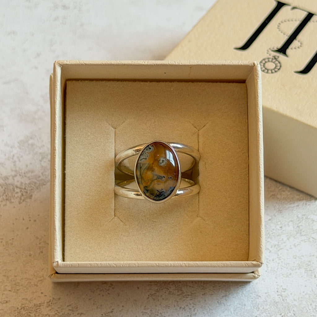 Handmade sterling silver double-band ring featuring an oval moss agate and carnelian gemstone with warm golden tones, soft green inclusions and a natural blue optical illusion. One-of-a-kind artisan statement ring by Journey Through Jewellery, photographed in a cream jewellery 