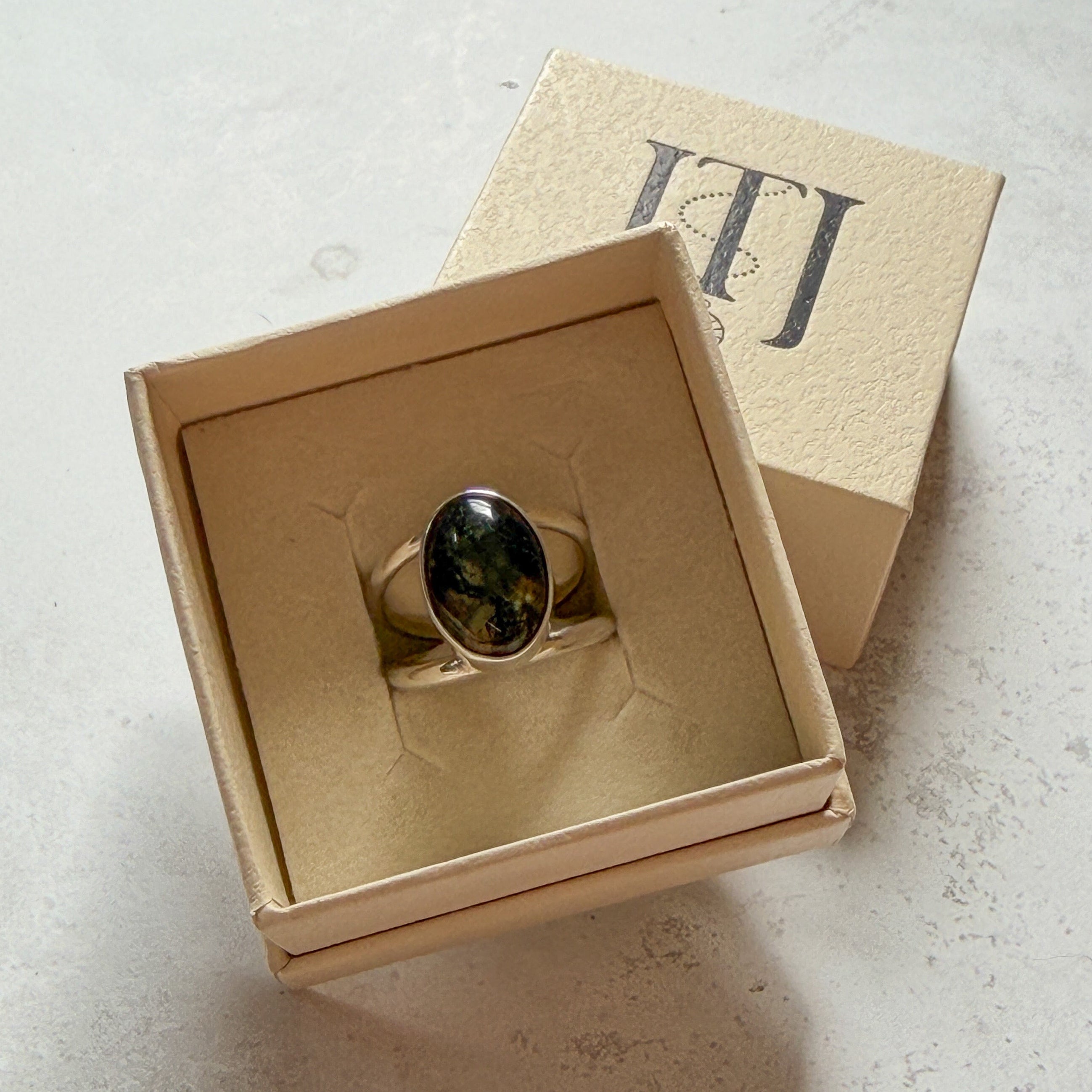 Handmade sterling silver double band ring featuring a one-of-a-kind oval moss agate gemstone with deep forest green inclusions. Contemporary artisan statement ring by Journey Through Jewellery, presented in a cream jewellery box with a mirror-polished finish.