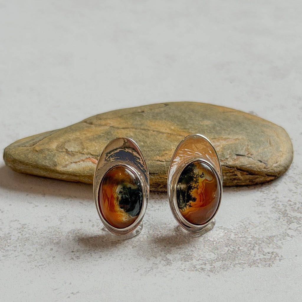 One-of-a-kind handcrafted sterling silver stud earrings featuring matching oval moss agate and carnelian gemstones with warm amber, deep green and natural landscape-like inclusions. Mirror-polished artisan earrings displayed on natural stone, with subtle variations making each gemstone unique