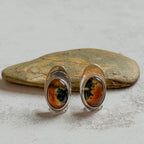 One-of-a-kind handcrafted sterling silver stud earrings featuring matching oval moss agate and carnelian gemstones with warm amber, deep green and natural landscape-like inclusions. Mirror-polished artisan earrings displayed on natural stone, with subtle variations making each gemstone unique