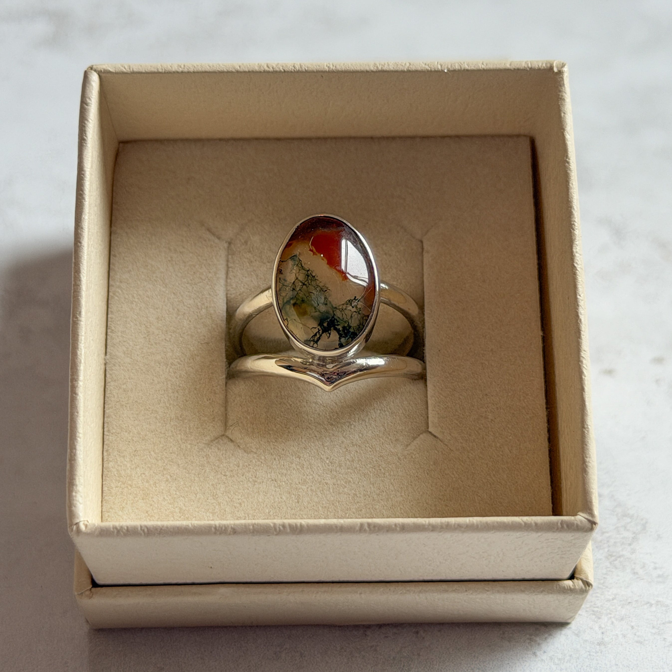 Pair of handcrafted sterling silver stud earrings with oval moss agate and carnelian gemstones, displayed in a cream Journey Through Jewellery gift box. Each one-of-a-kind gemstone features unique amber, green and black landscape-like inclusions, finished with mirror-polished bezels and handcrafted silver grain details.