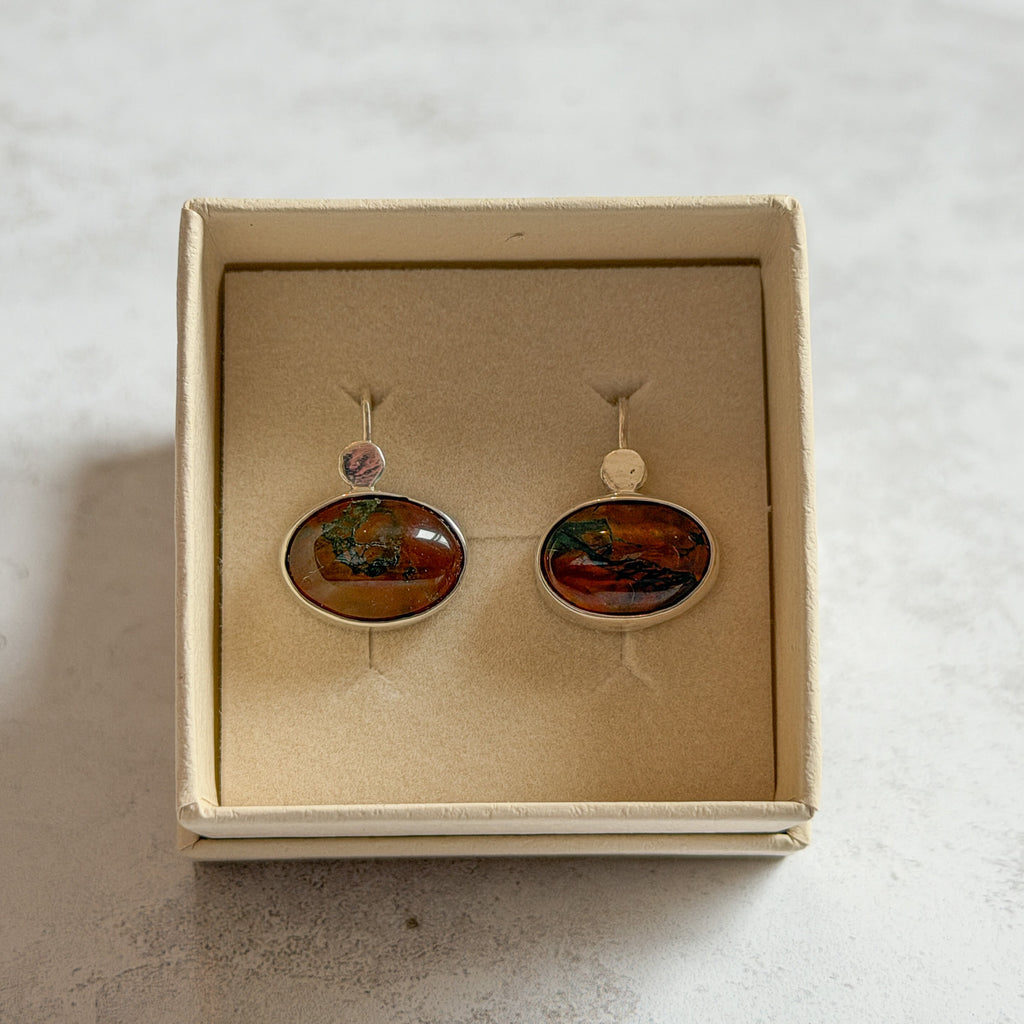 Pair of handcrafted sterling silver stud earrings with oval moss agate and carnelian gemstones, displayed in a cream Journey Through Jewellery gift box. Each one-of-a-kind gemstone features unique amber, green and black landscape-like inclusions, finished with mirror-polished bezels and handcrafted silver grain details.