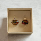 Pair of handcrafted sterling silver stud earrings with oval moss agate and carnelian gemstones, displayed in a cream Journey Through Jewellery gift box. Each one-of-a-kind gemstone features unique amber, green and black landscape-like inclusions, finished with mirror-polished bezels and handcrafted silver grain details.