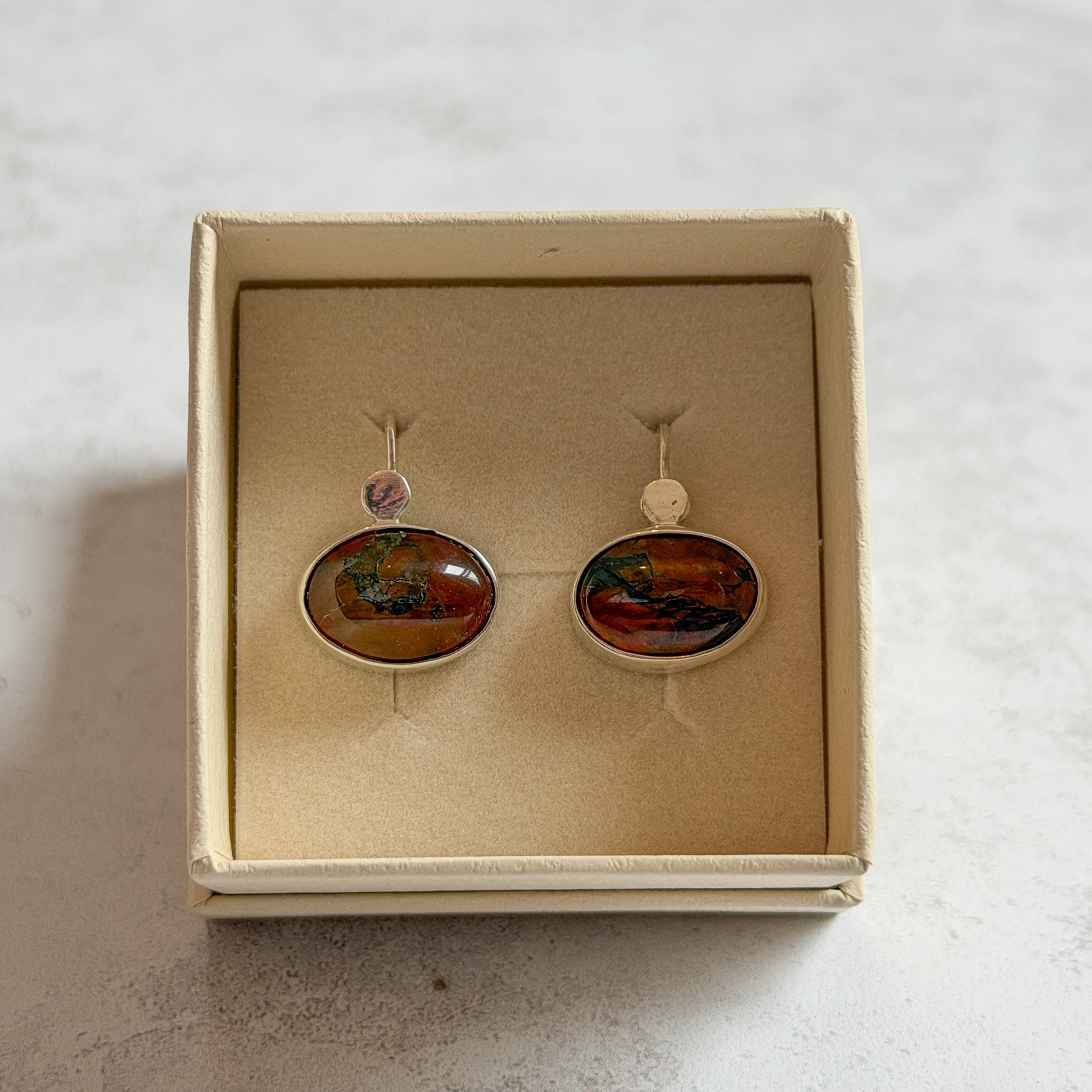 Pair of handcrafted sterling silver stud earrings with oval moss agate and carnelian gemstones, displayed in a cream Journey Through Jewellery gift box. Each one-of-a-kind gemstone features unique amber, green and black landscape-like inclusions, finished with mirror-polished bezels and handcrafted silver grain details.