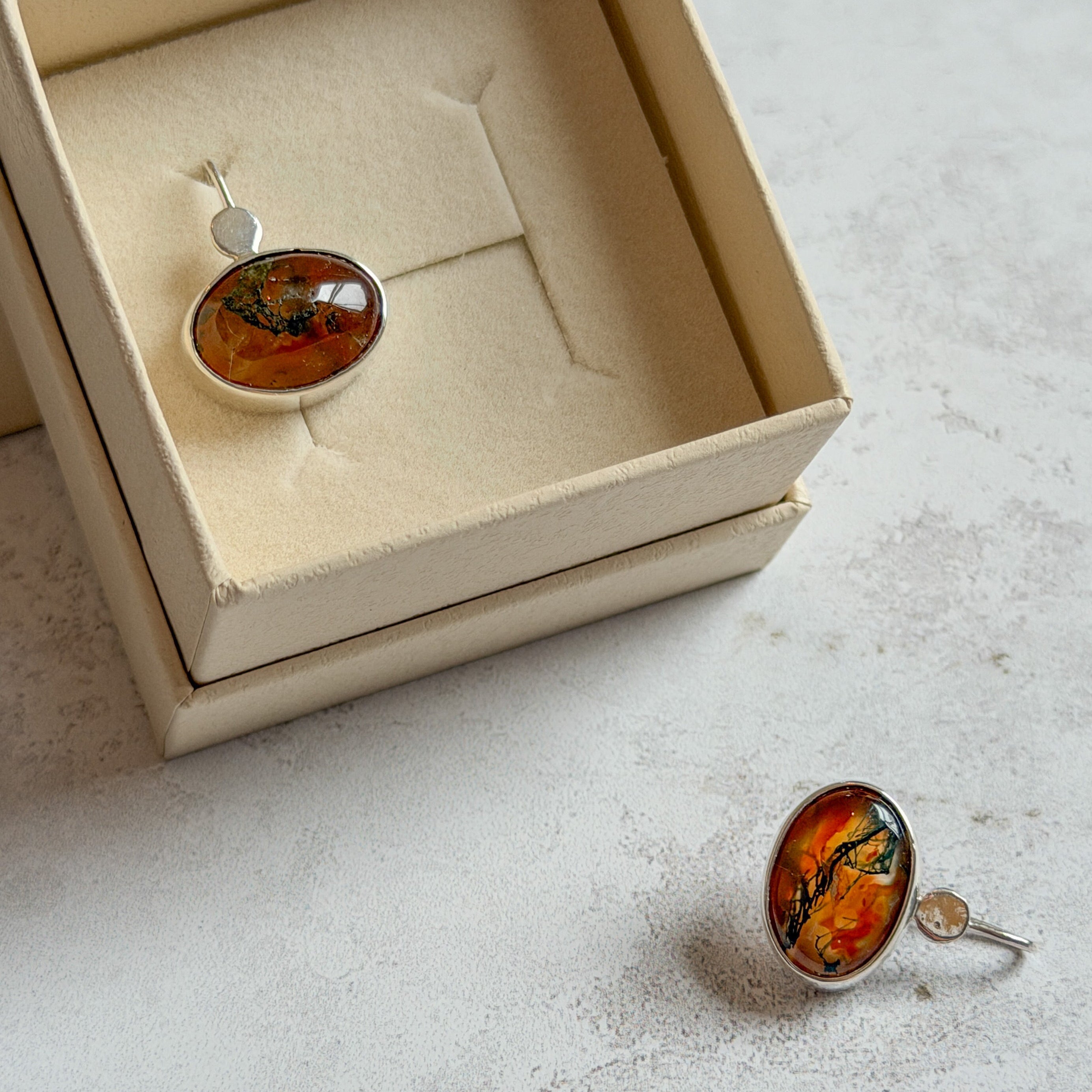 Handmade sterling silver stud earrings featuring oval moss agate and carnelian gemstones with rich amber, green and black natural inclusions. One earring is displayed in a cream Journey Through Jewellery gift box while the other rests beside it, showcasing one-of-a-kind artisan jewellery with mirror-polished bezels and signature open-back settings.