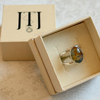 Handmade sterling silver double-band ring featuring an oval moss agate and carnelian gemstone with warm golden tones, soft green inclusions and a natural blue optical illusion. One-of-a-kind artisan statement ring by Journey Through Jewellery, photographed in a cream jewellery 