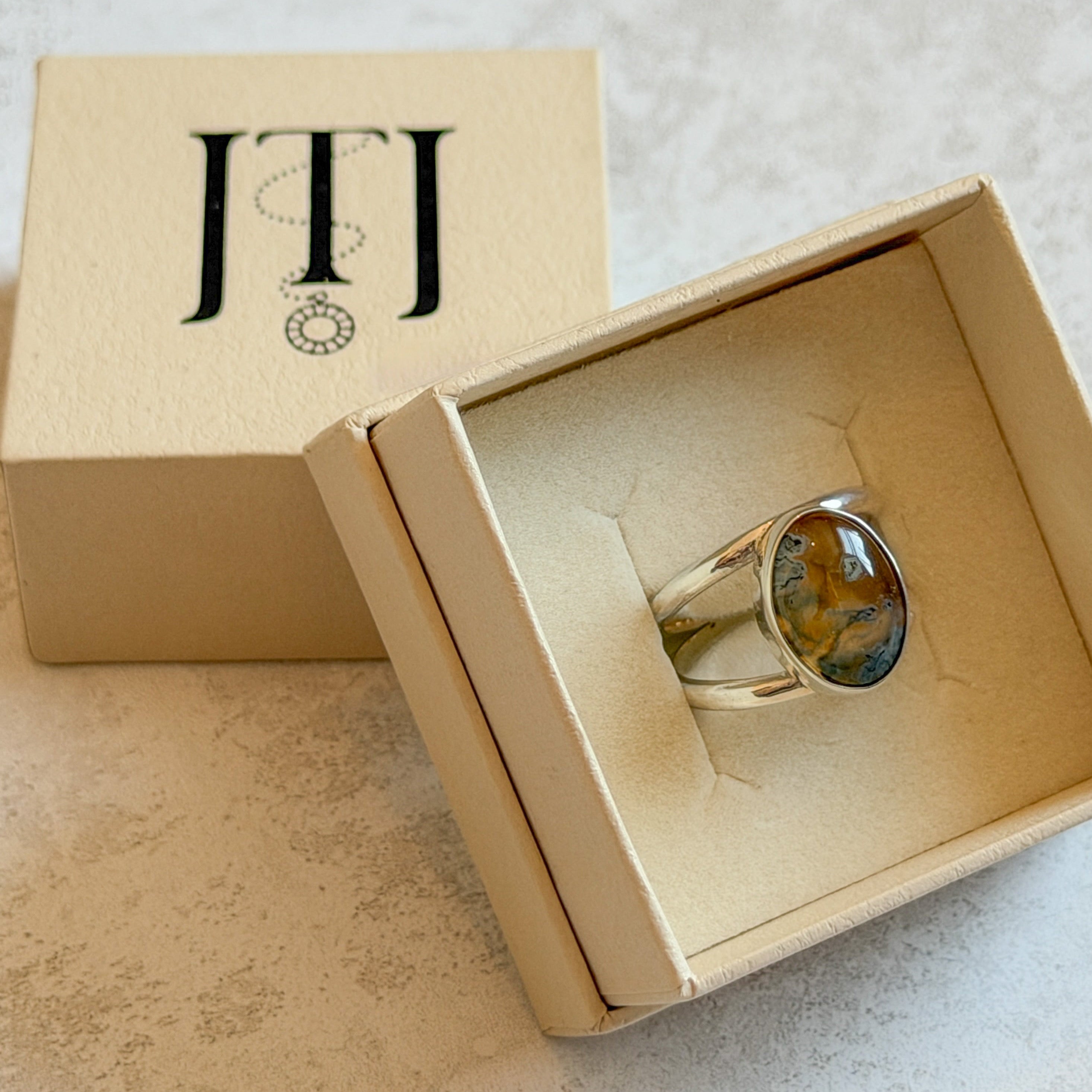 Handmade sterling silver double-band ring featuring an oval moss agate and carnelian gemstone with warm golden tones, soft green inclusions and a natural blue optical illusion. One-of-a-kind artisan statement ring by Journey Through Jewellery, photographed in a cream jewellery 