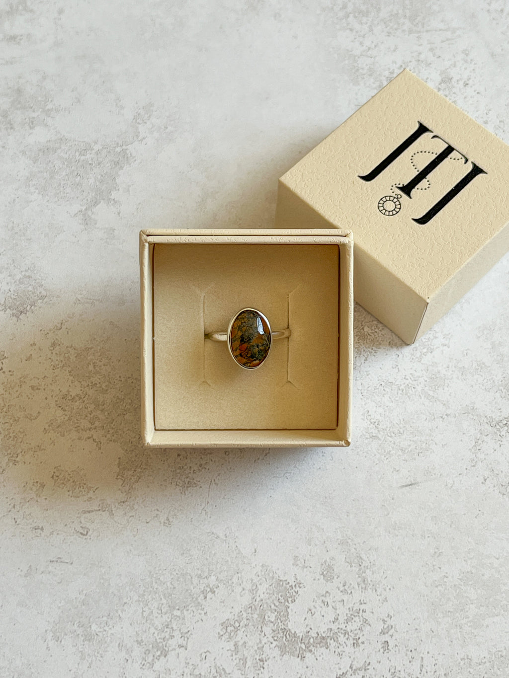 Handmade sterling silver ring with oval moss agate and warm carnelian tones, bezel set on a D wire band with organic cut-out window, displayed in a cream jewellery box on a neutral textured background