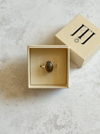 Handmade sterling silver ring with oval moss agate and warm carnelian tones, bezel set on a D wire band with organic cut-out window, displayed in a cream jewellery box on a neutral textured background