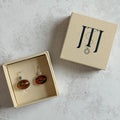 Pair of handcrafted sterling silver stud earrings with oval moss agate and carnelian gemstones, displayed in a cream Journey Through Jewellery gift box. Each one-of-a-kind gemstone features unique amber, green and black landscape-like inclusions, finished with mirror-polished bezels and handcrafted silver grain details.