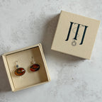 Pair of handcrafted sterling silver stud earrings with oval moss agate and carnelian gemstones, displayed in a cream Journey Through Jewellery gift box. Each one-of-a-kind gemstone features unique amber, green and black landscape-like inclusions, finished with mirror-polished bezels and handcrafted silver grain details.