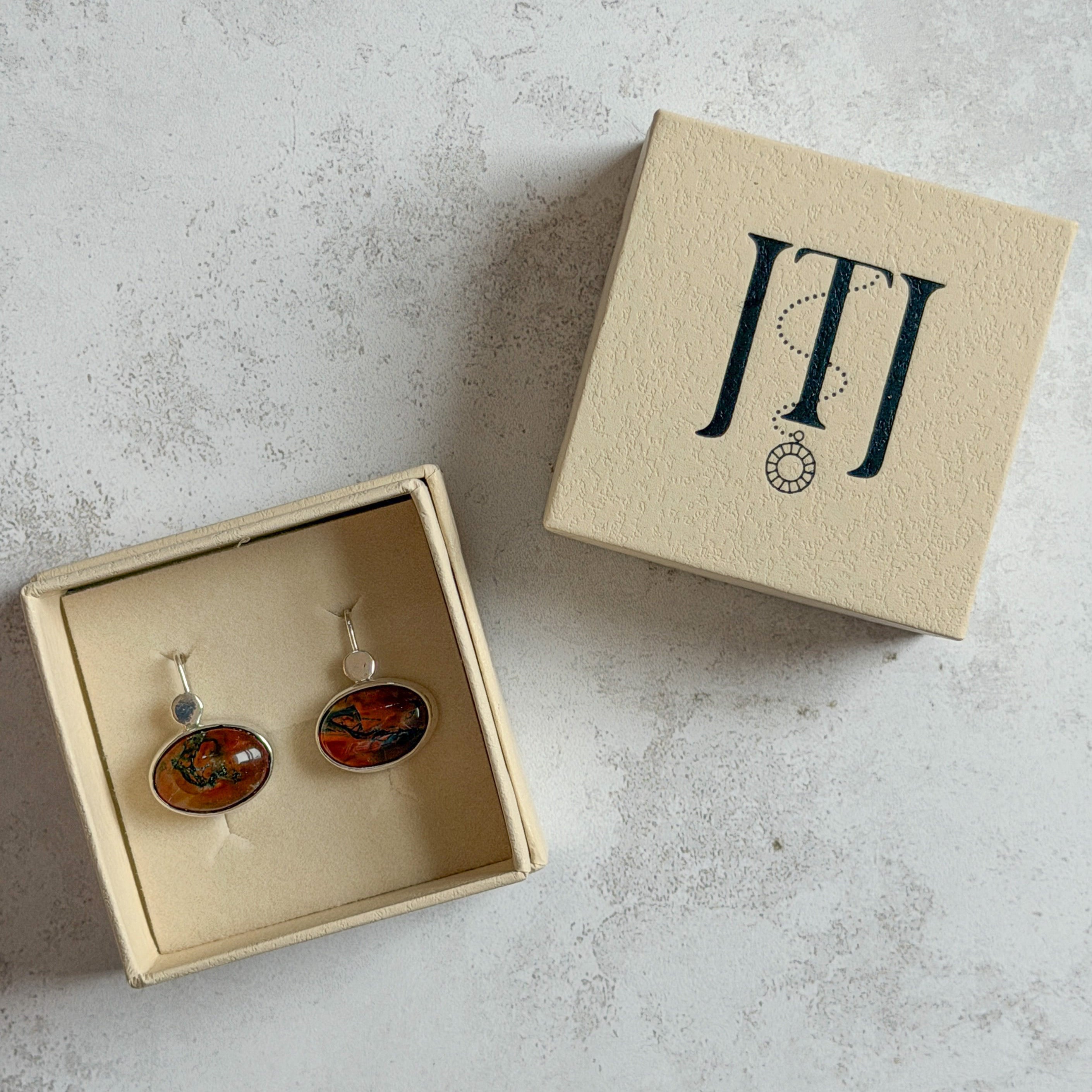 Pair of handcrafted sterling silver stud earrings with oval moss agate and carnelian gemstones, displayed in a cream Journey Through Jewellery gift box. Each one-of-a-kind gemstone features unique amber, green and black landscape-like inclusions, finished with mirror-polished bezels and handcrafted silver grain details.
