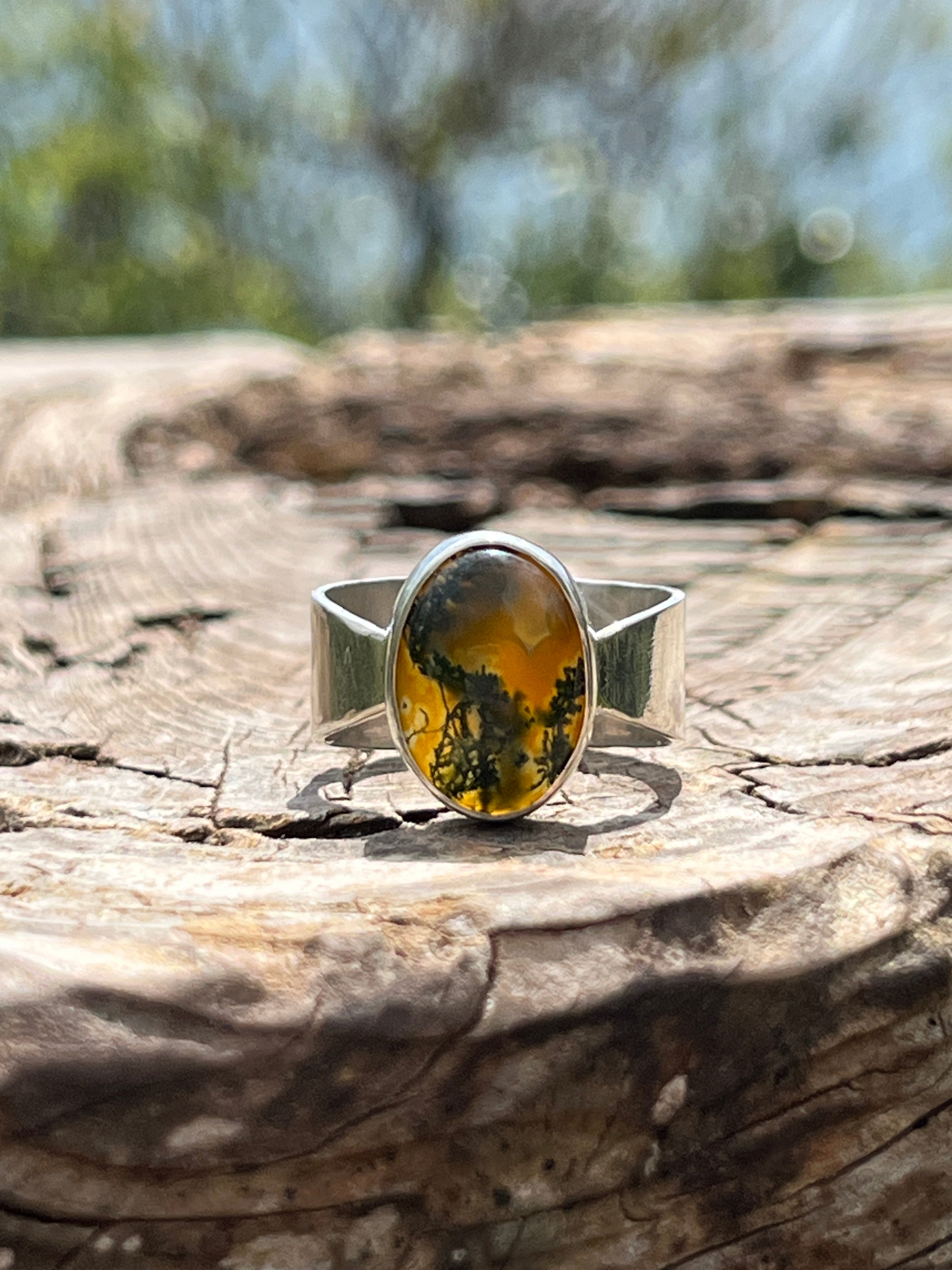 Handmade sterling silver ring with oval moss agate and carnelian gemstone, bezel set on a wide tapered silver band, photographed outdoors on textured wood in natural light.