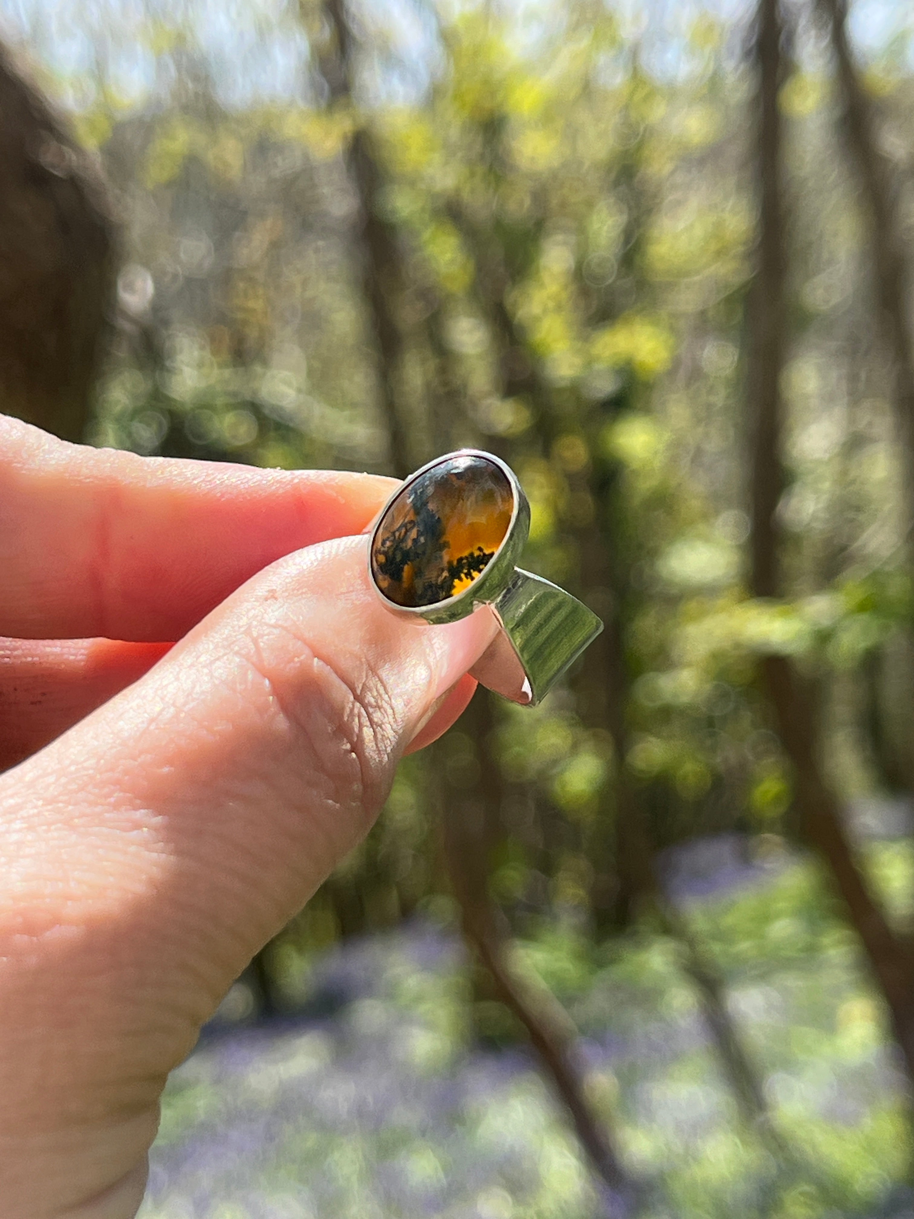 Handmade sterling silver ring with oval moss agate and carnelian gemstone, bezel set on a  tapered wide band, held in hand against a soft woodland background with natural light shining through the stone.